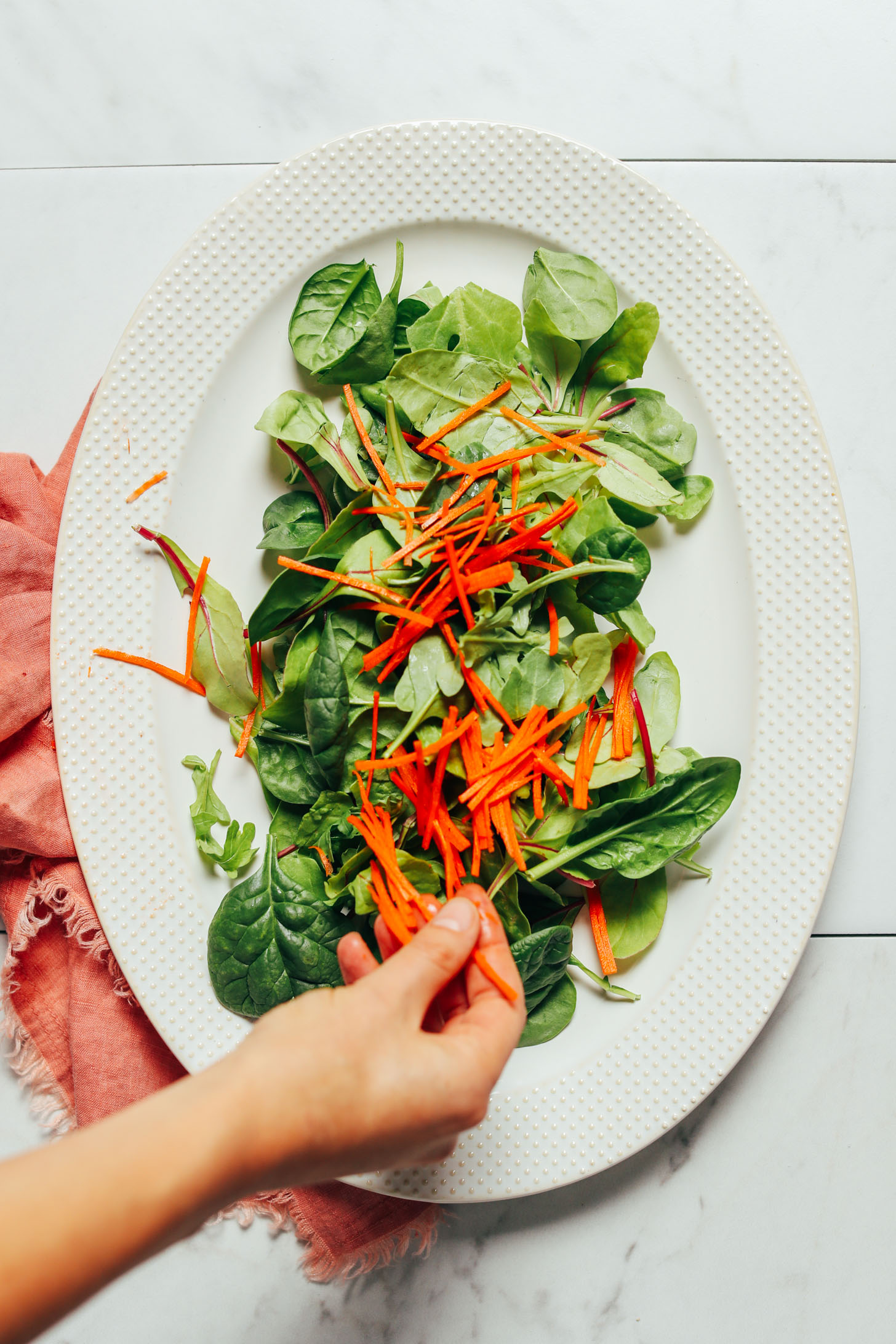 Overhead image of white plate with greens a pink towel underneath and carrots being placed on top by hand