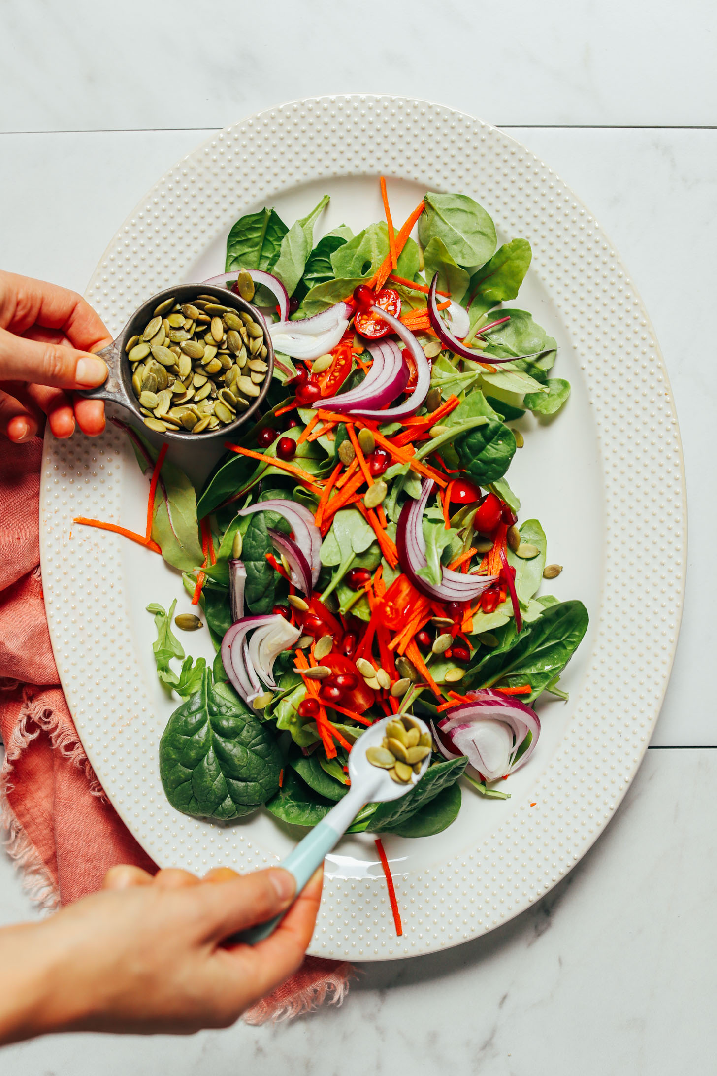 Overhead image of salad with greens carrots and onion and a measuring cup with pumpkin seeds with a hand sprinkling some seeds on top