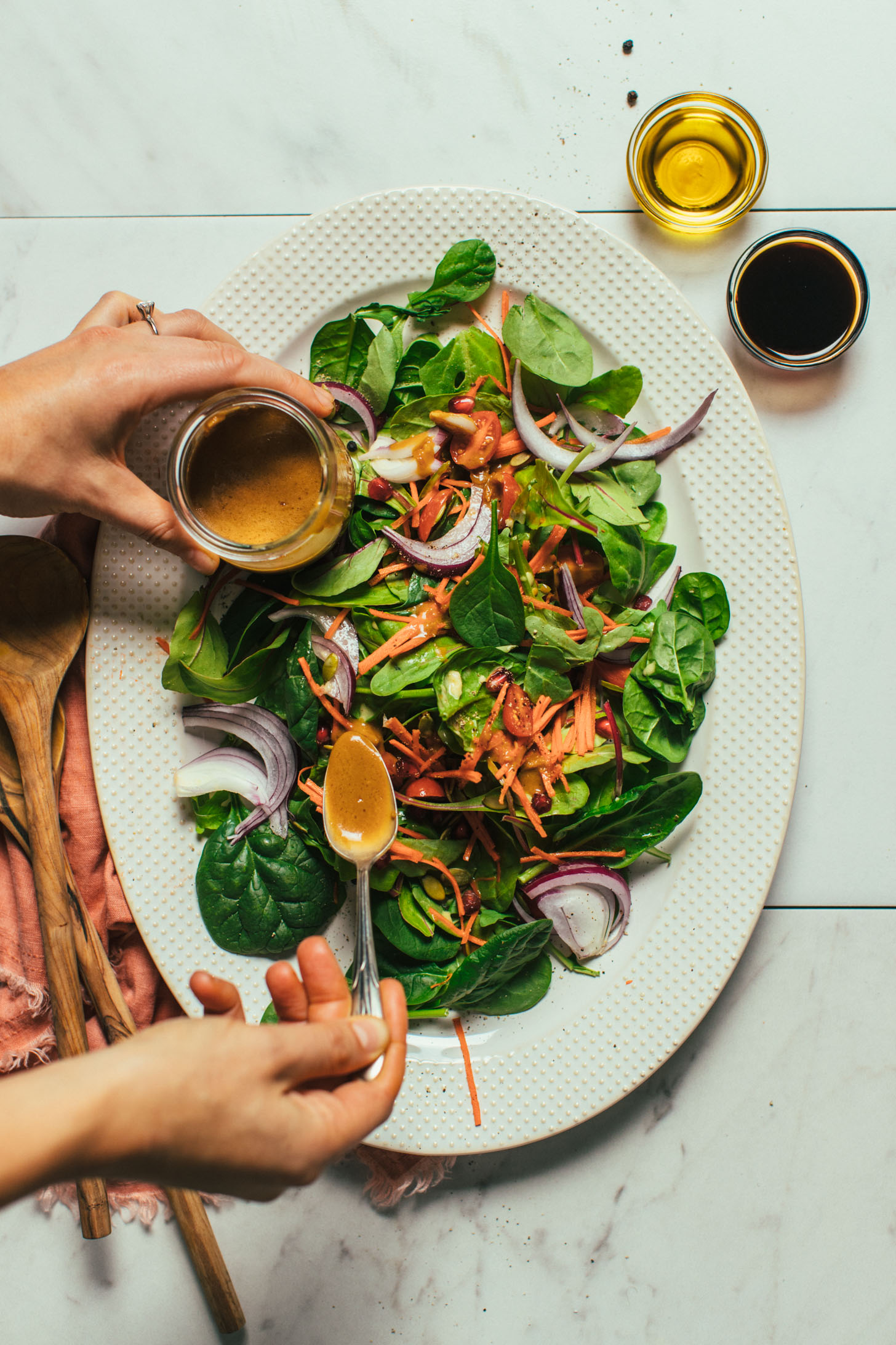 Holding a jar of dressing in one hand and using a spoon in the other hand to drizzle balsamic dressing on a salad
