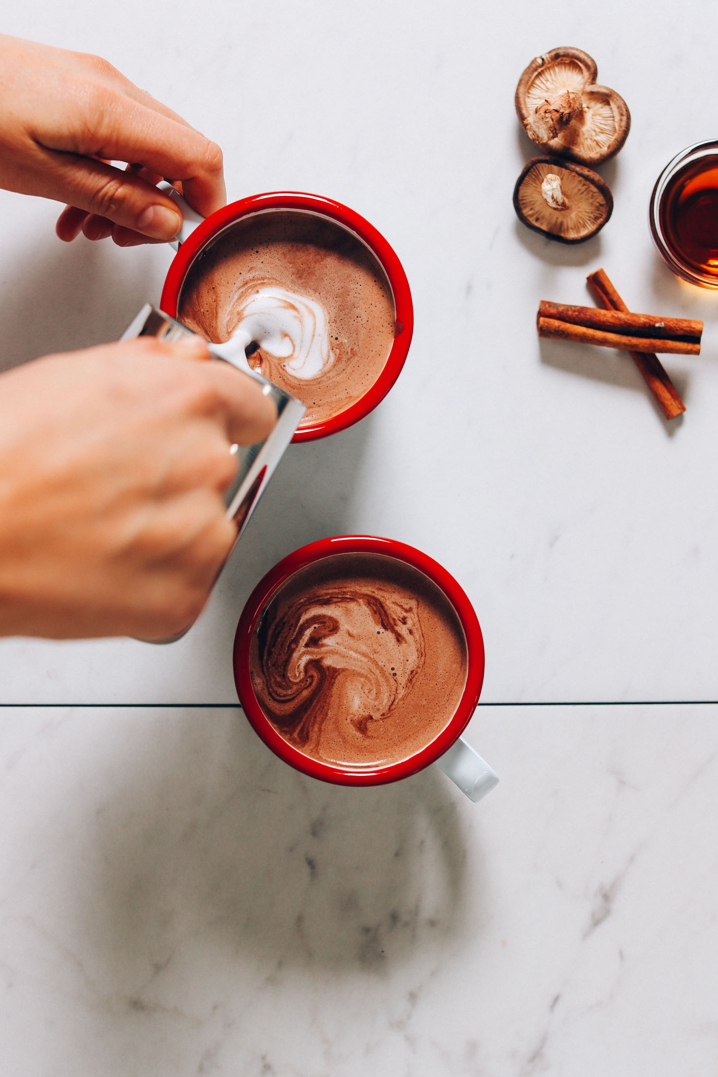 Overhead image of hot cacao mushroom latte being topped with steamed milk, with mushrooms and cinnamon sticks in the top right corner