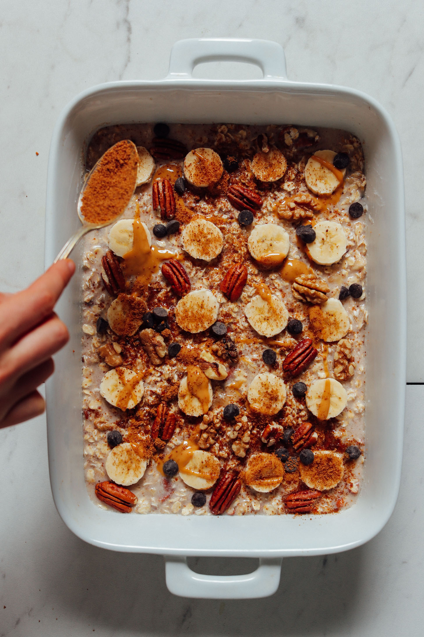 Overhead image of banana oatmeal in a baking dish lined with ripe banana being topped with a spoonful of coconut sugar