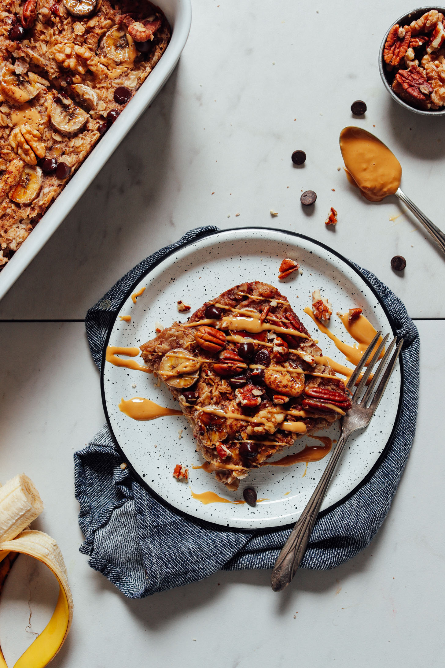 Image of a serving of banana baked oatmeal on a white plate, garnished with nut butter and nuts, chocolate chips, and banana on the side