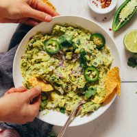 Overhead image of guacamole in a white bowl and a hand scooping some guacamole with a chip