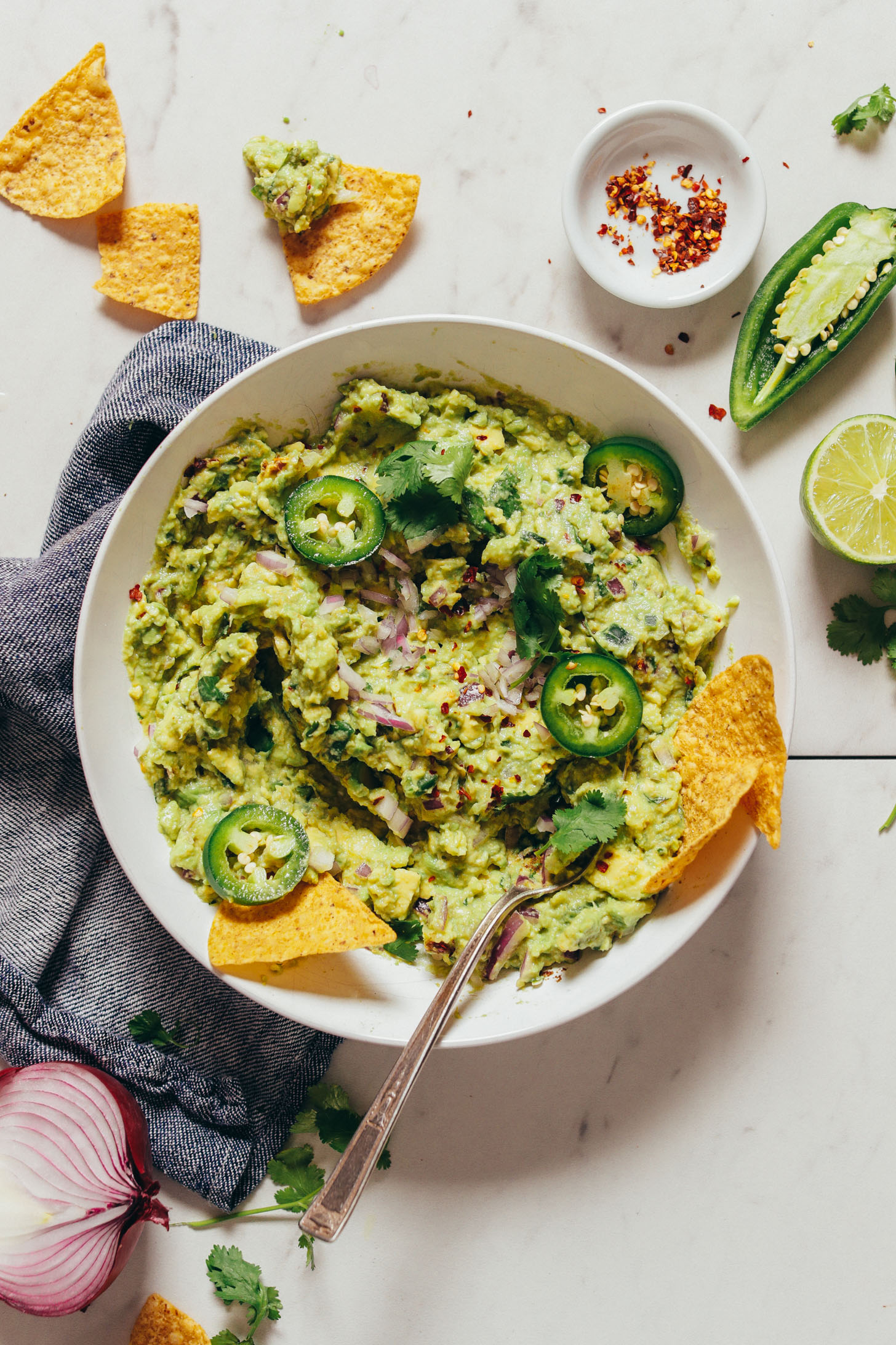 Overhead image of guacamole in a white bowl over a blue towel, garnished with jalapenos, red onion, and cilantro