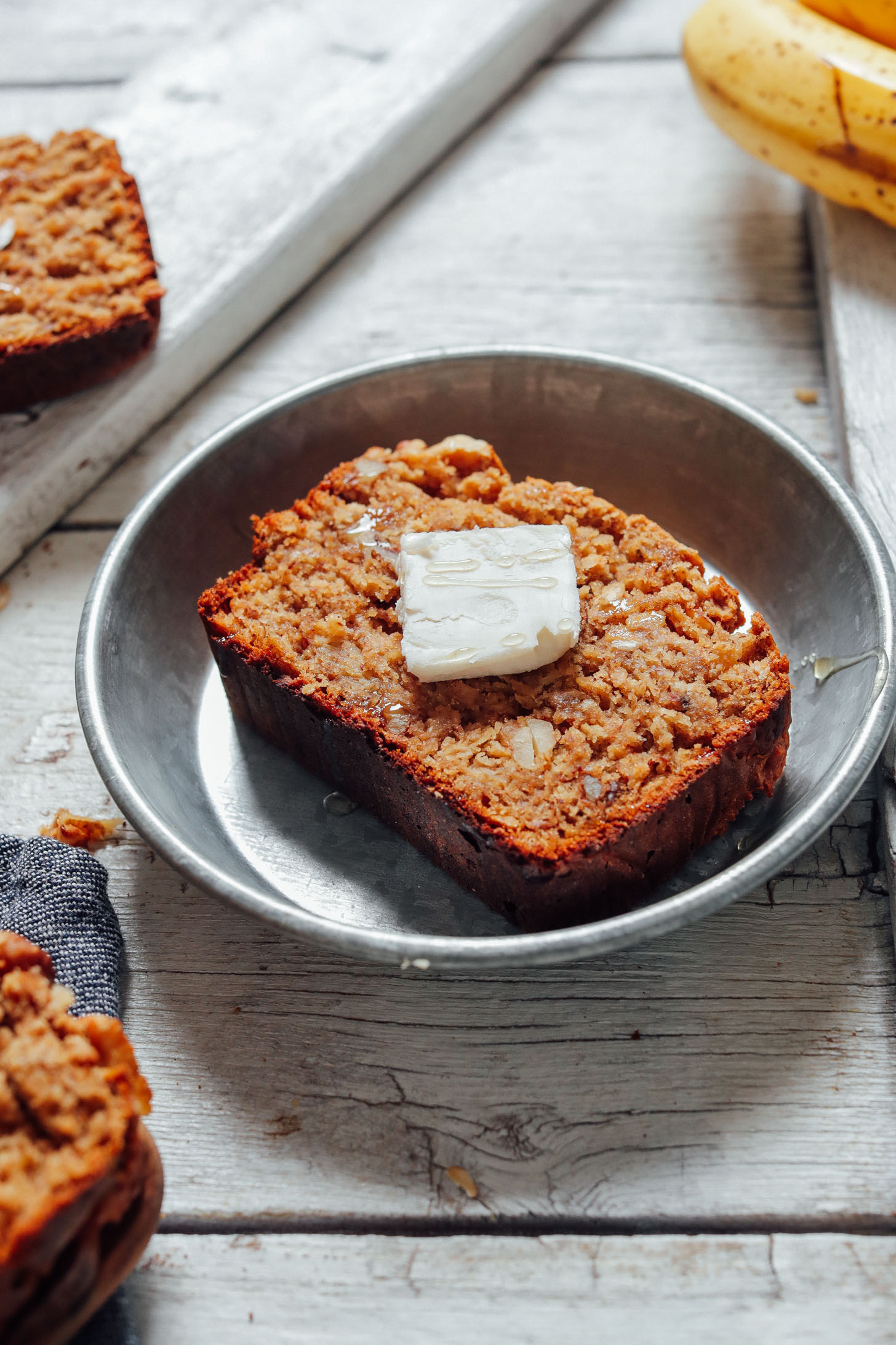 Image of vegan gluten free banana bread slice on a metal plate with vegan butter on top and bananas in the background