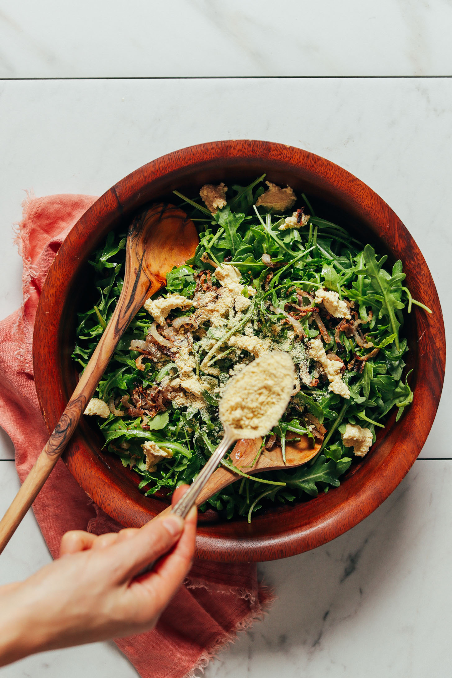 Sprinkling cashew parmesan cheese onto a big bowl of our Lemony Arugula Salad with Crispy Shallot recipe