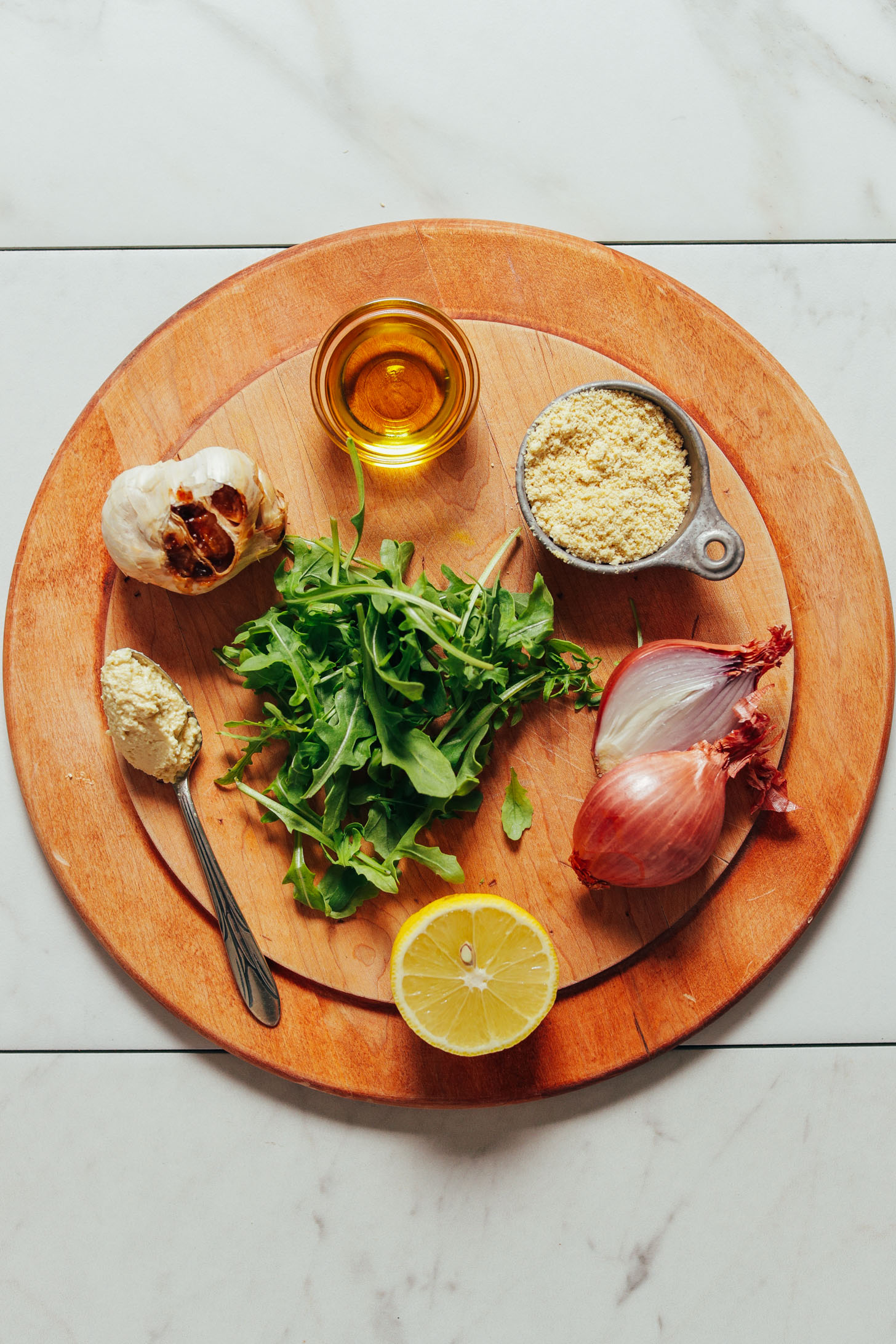 Wood cutting board with arugula, shallots, garlic, lemon, and other ingredients to make our simple Lemony Arugula Salad with Crispy Shallot recipe