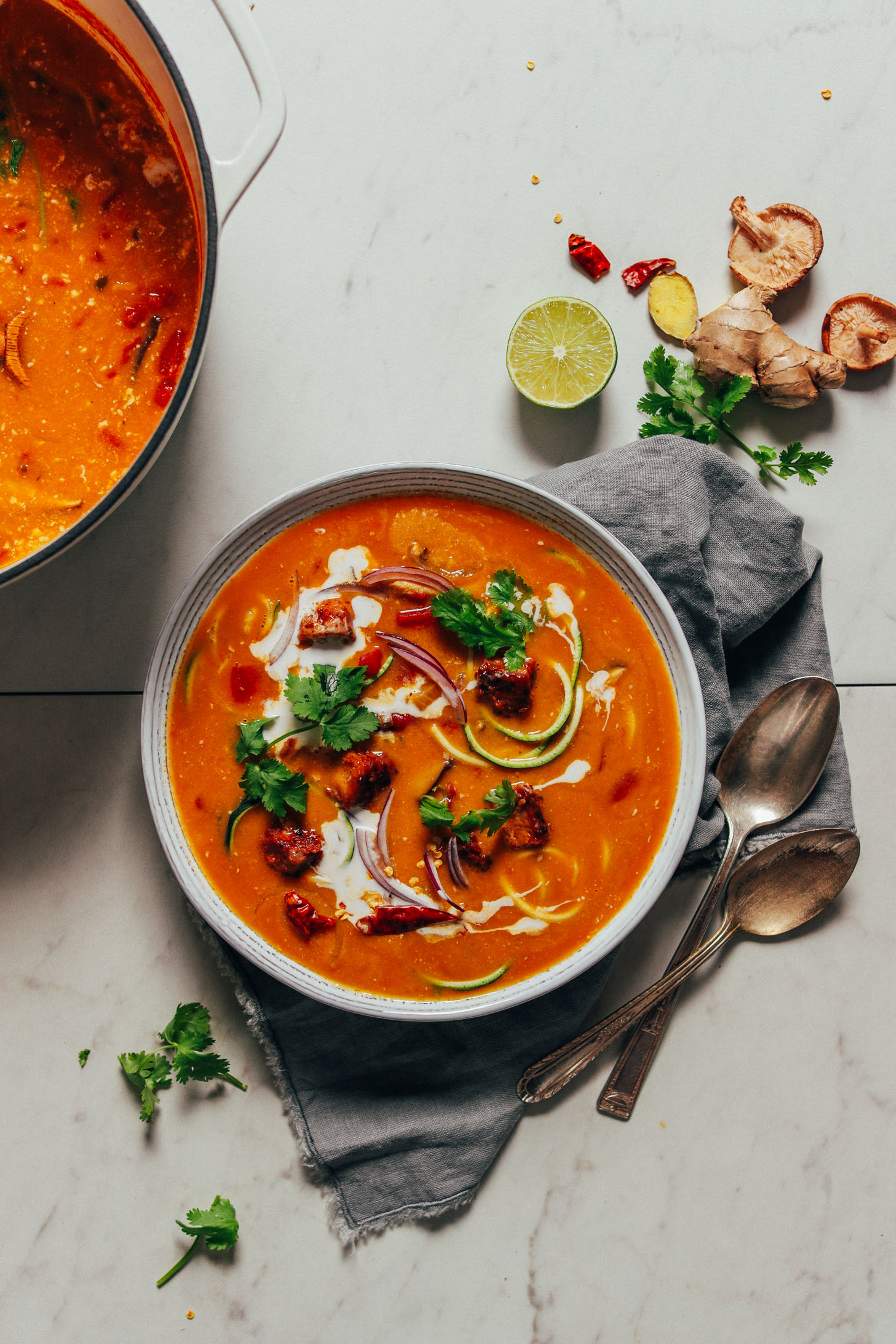 Bowl and pot of Vegan Tom Yum Soup alongside ginger, lime, and other ingredients used to make it