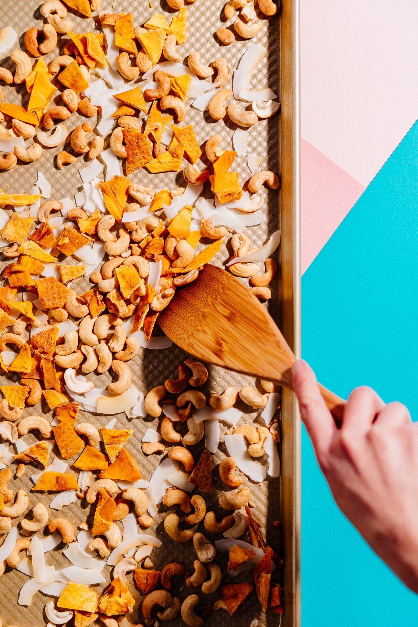 Using a wood spatula to mix healthy homemade Tropical Trail Mix on a baking sheet