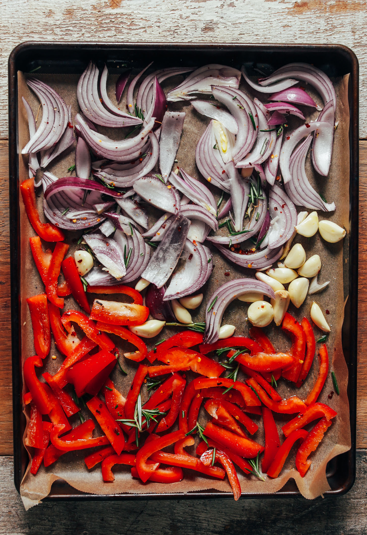 Parchment-lined baking sheet with peeled garlic, red onion, red bell pepper, and rosemary ready to be roasted