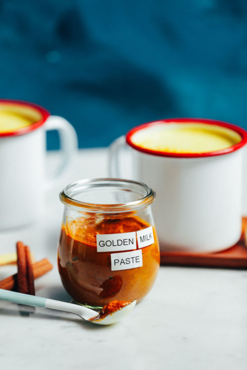 Jar of golden milk mix with two mugs of golden milk behind it