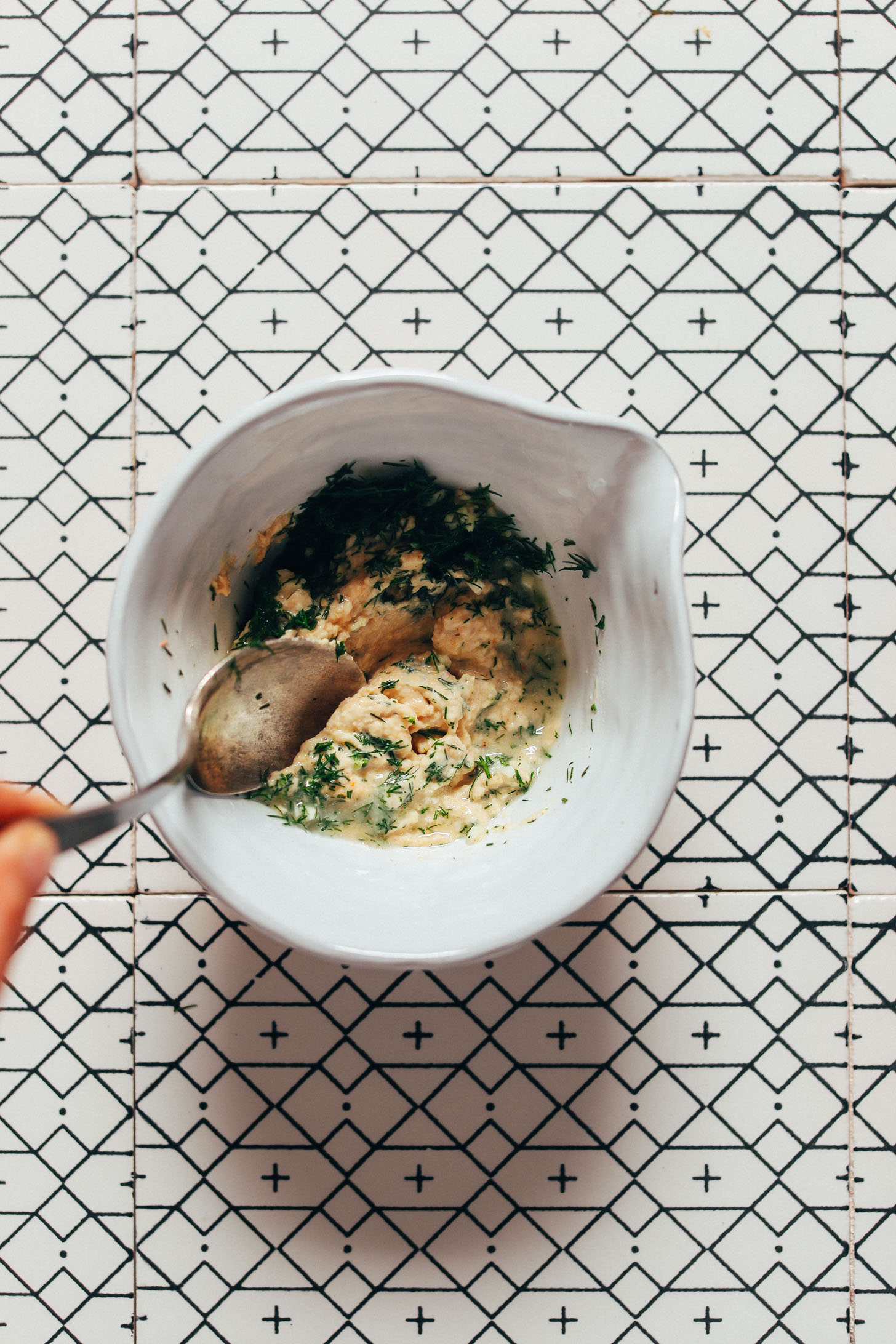 Spoon mixing hummus, lemon juice, minced garlic, and fresh dill in a small ceramic bowl for garlic dairy-free dill sauce