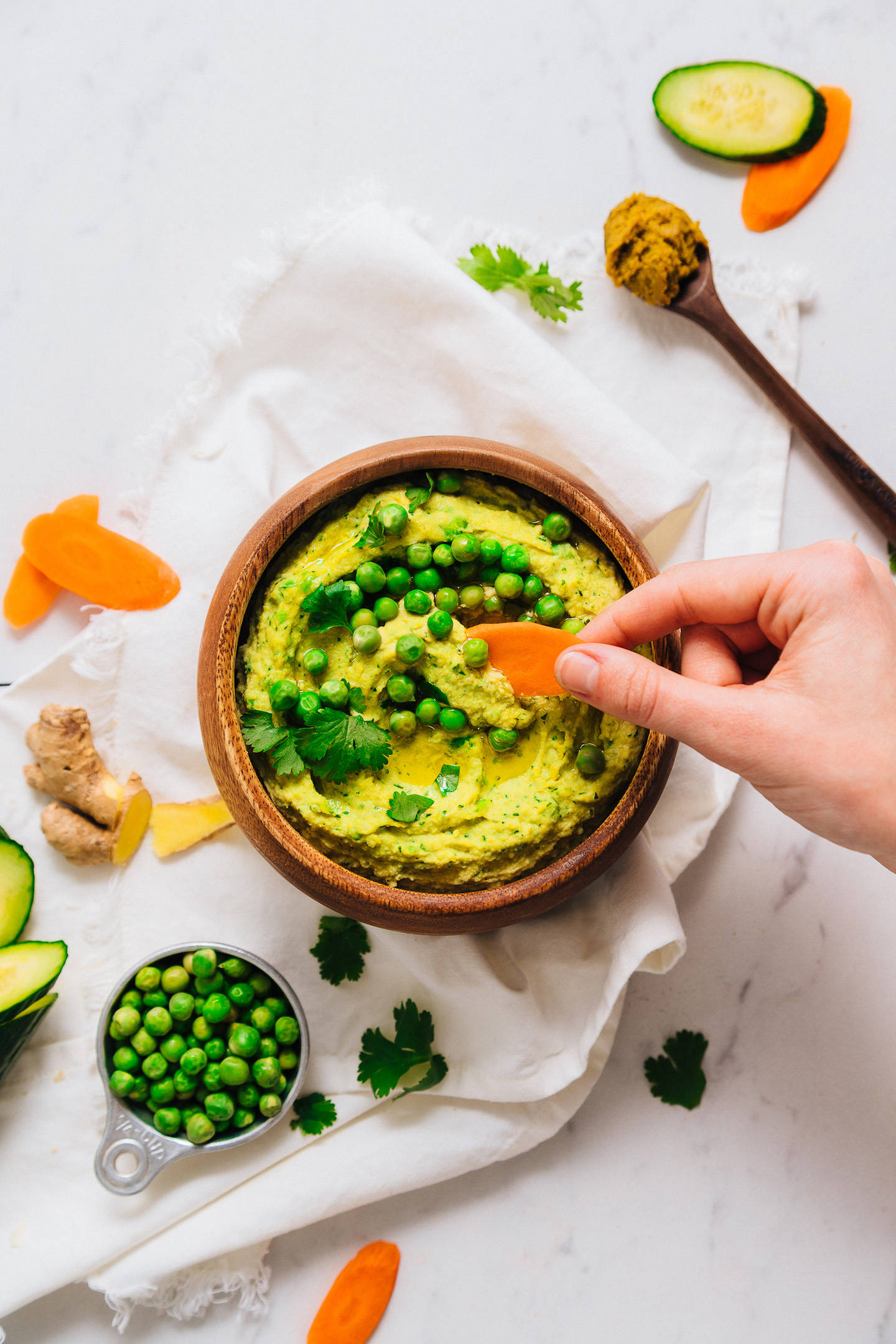 Using a carrot slice to scoop up a bite of Green Pea Curry Hummus from a wood bowl