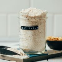 Jar and scoop of homemade oat flour sitting beside a bowl of oats