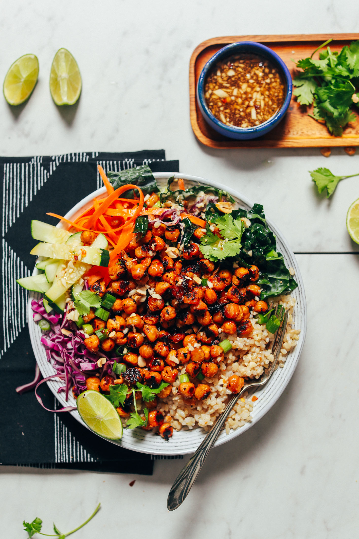 Bowl of brown rice, fresh vegetables, Miso Chickpeas, cilantro, and lime wedges surrounded by more ingredients for serving
