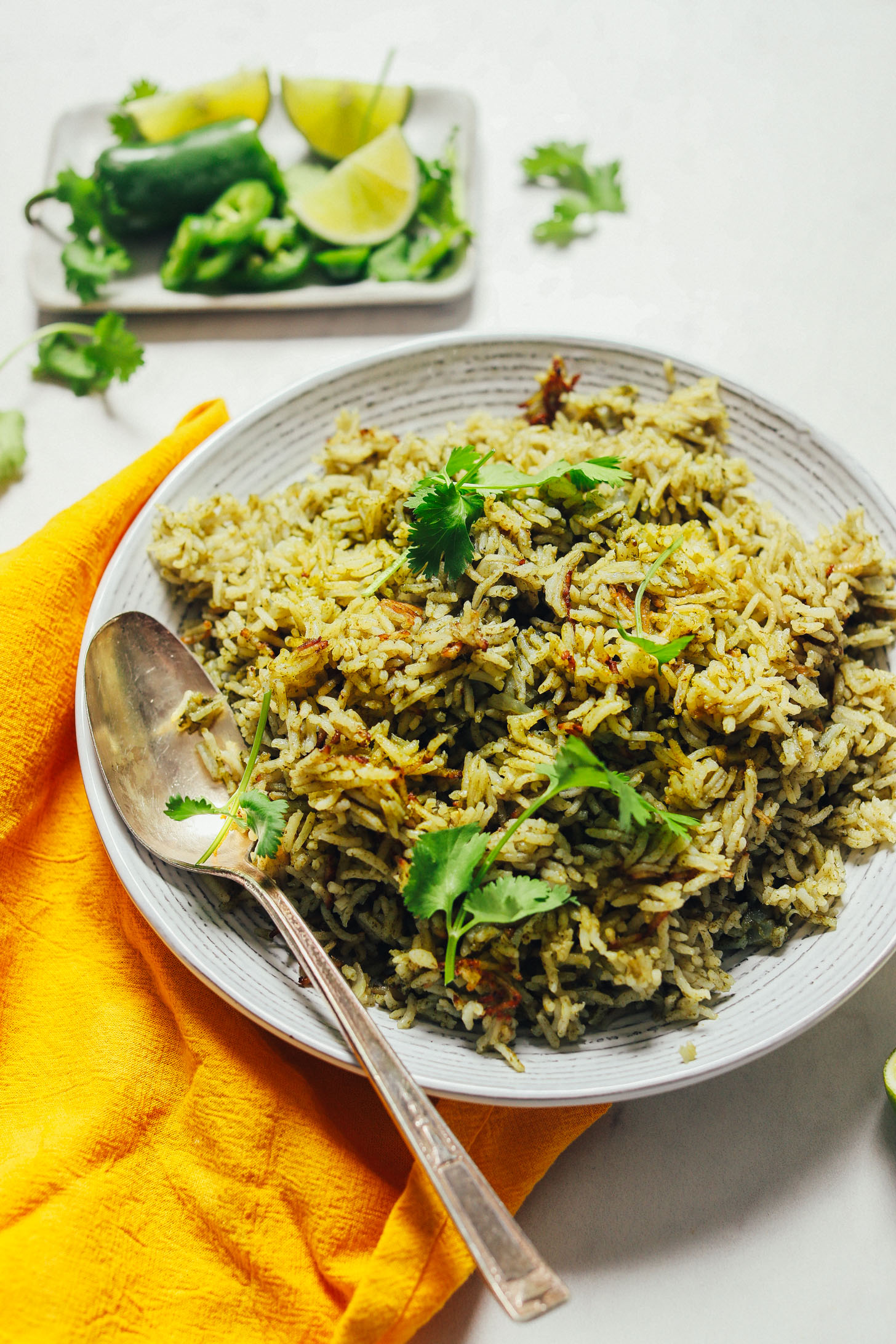 Spoon resting in a bowl of gluten-free Green Rice beside fresh limes, jalapeno, and cilantro