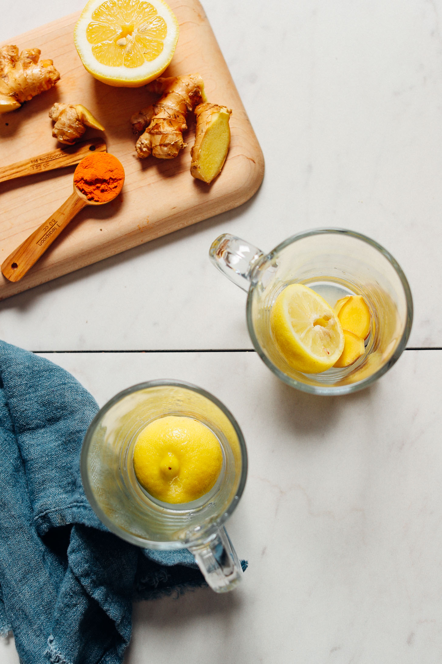 overhead image of two clear mugs with lemon and ginger, a blue towel to the left, and a cutting board above with lemon, ginger, and spices