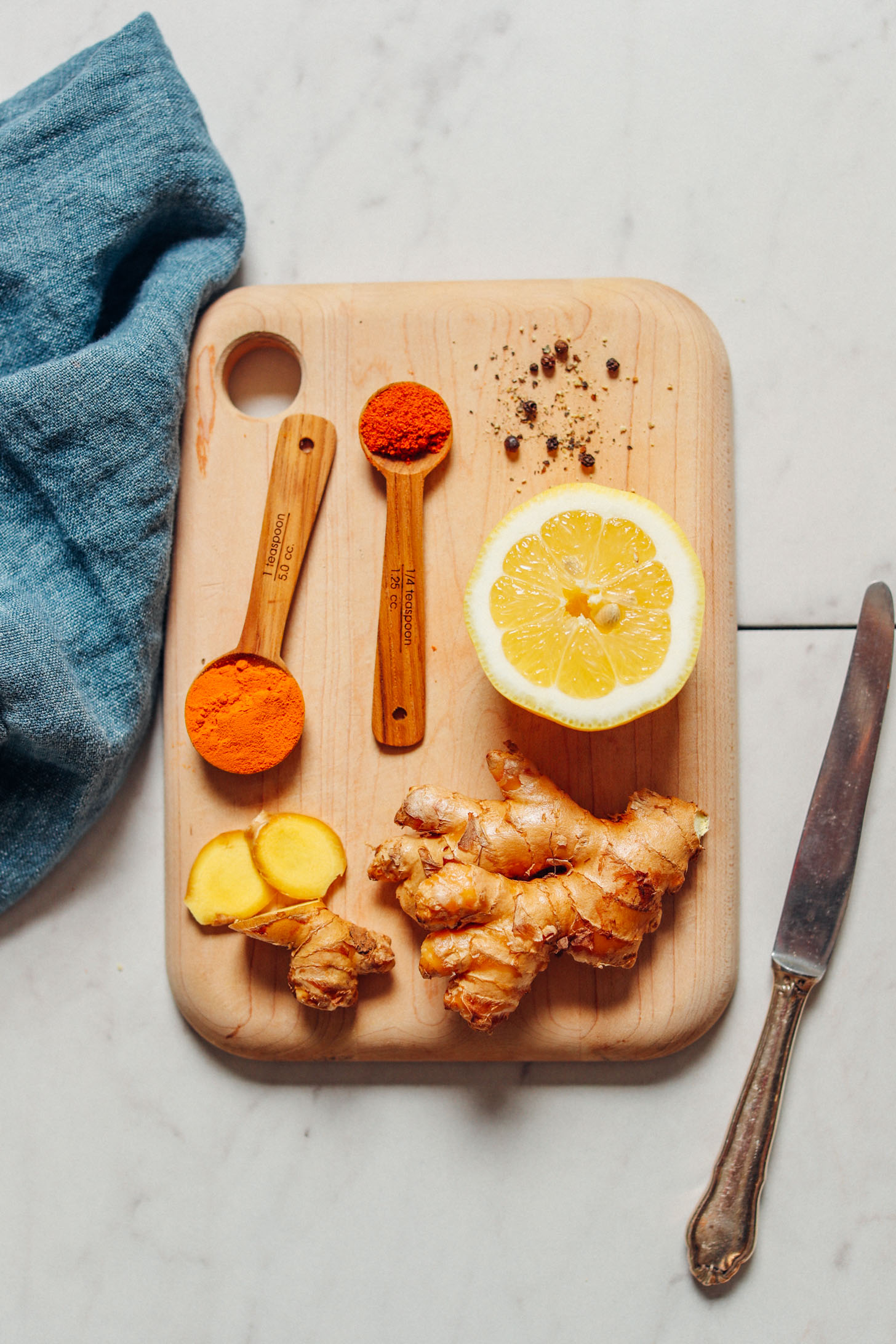 image of lemon ginger water ingredients from overhead with lemon ginger turmeric and cayenne on a cutting board a blue cloth beside it and a knife to the right