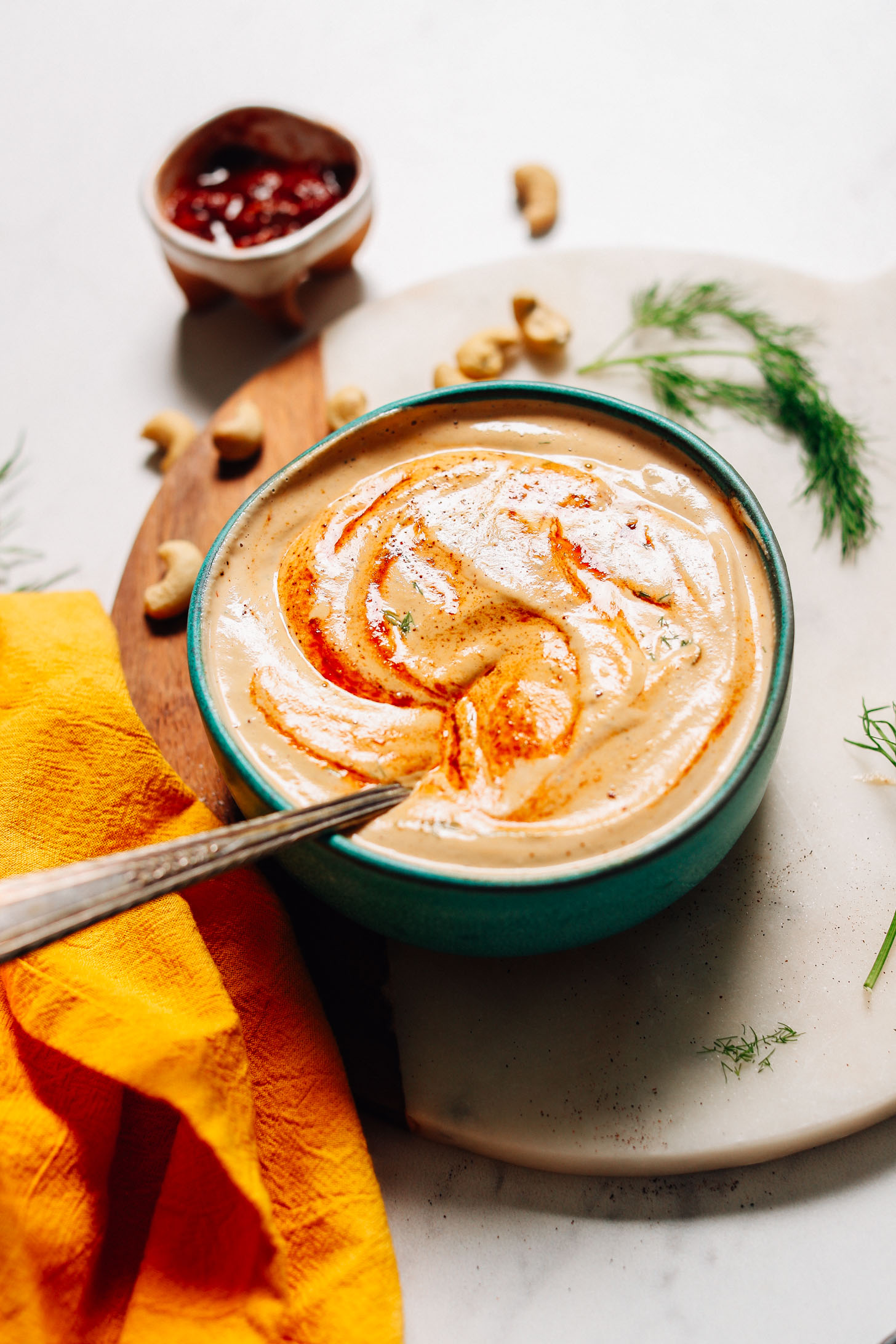 Spoon resting in a bowl of Vegan Chipotle Ranch Dressing surrounded by cashews, fresh dill, and chipotle peppers in adobo
