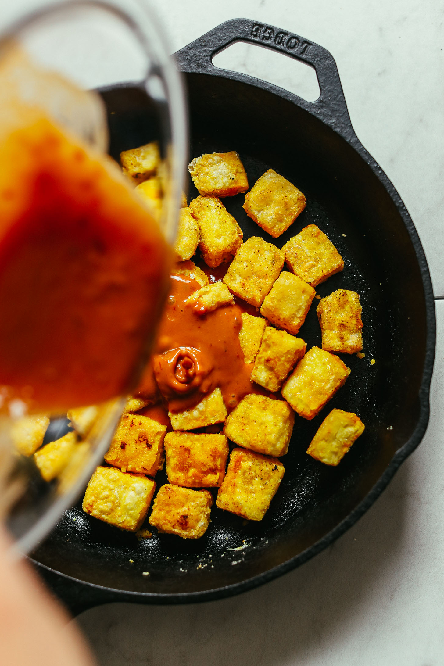 overhead image of cornstarch-coated tofu being cooked in a cast iron skillet and peanut sauce being poured on top