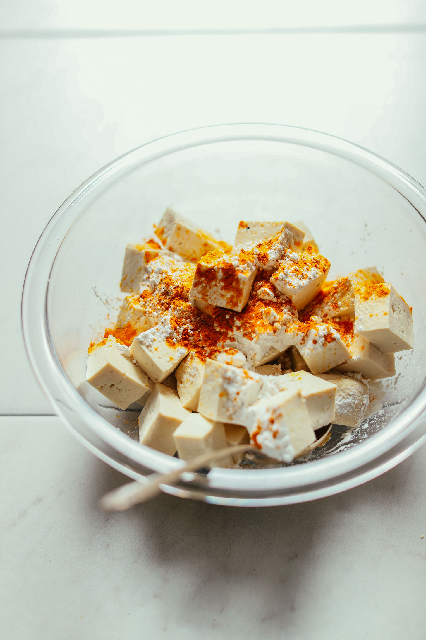 Cubed tofu seasoned with curry powder and cornstarch, tossed with a metal spoon in a glass bowl