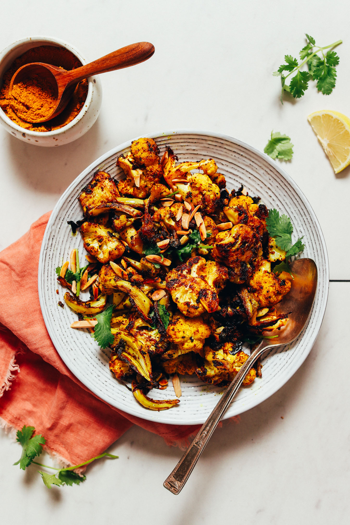Bowl of Quick Crispy Cauliflower topped with toasted almonds and fresh cilantro beside a dish of curry powder