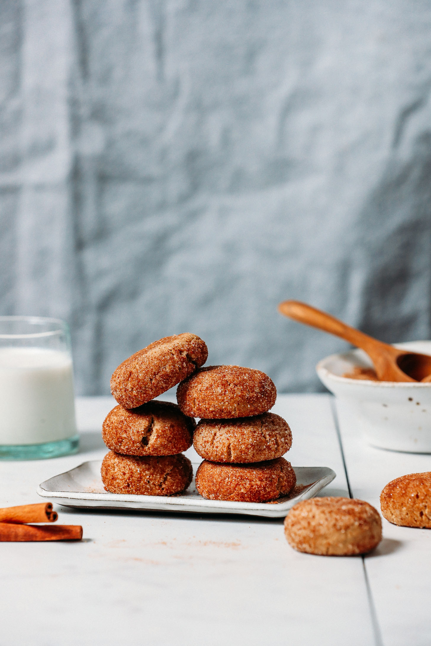 GF Vegan Snickerdoodle Cookies piled on a plate next to a glass of non-dairy milk, a cinnamon stick, and cinnamon sugar coating
