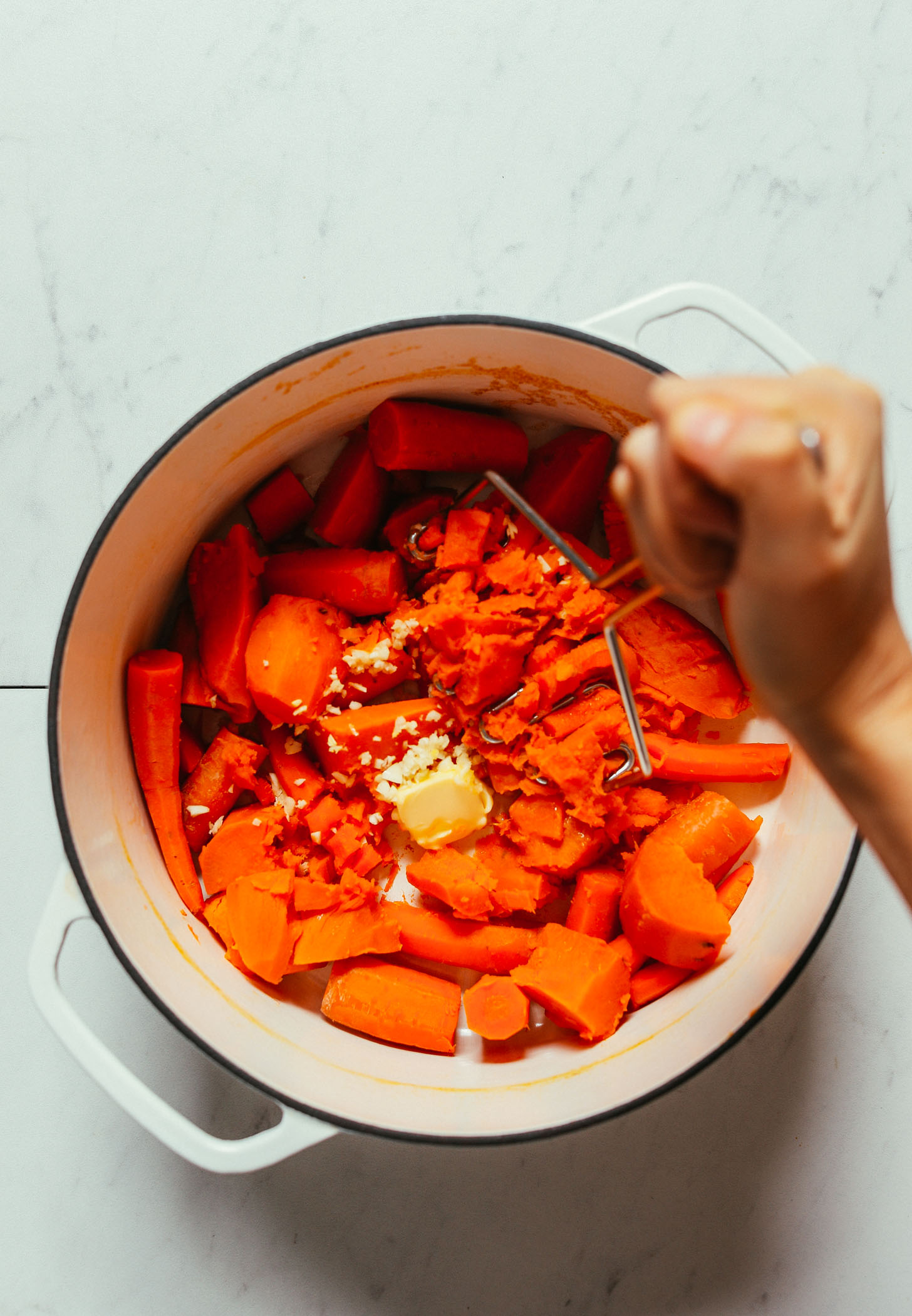 Using a potato masher to mash sweet potatoes and carrots with vegan butter and minced garlic