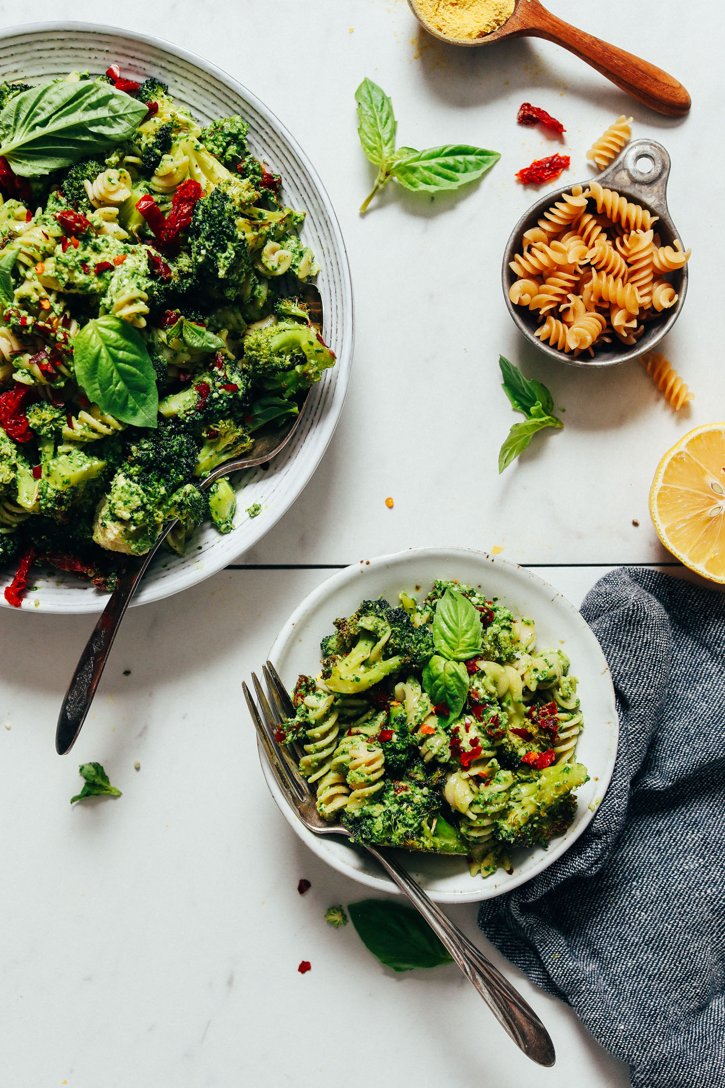 Bowls of Pesto Broccoli Salad beside dry pasta, lemon juice, and sun-dried tomatoes