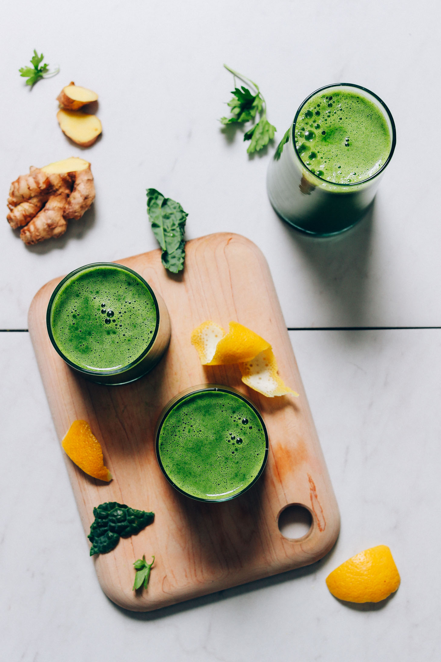 Glasses of homemade Green Juice on a cutting board beside ingredients used to make it
