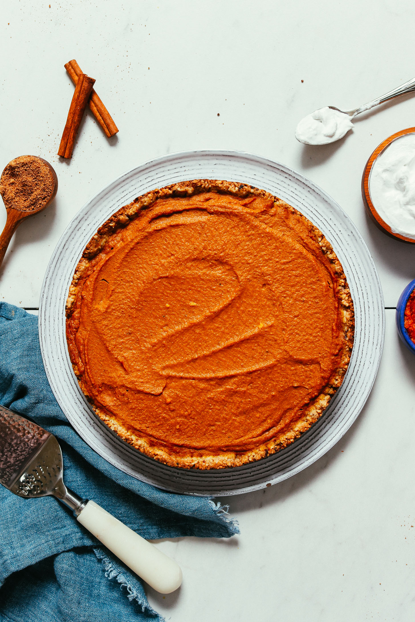 Vegan gluten-free Pumpkin Pie on a plate surrounded by cinnamon sticks, coconut sugar, pumpkin purée, and coconut whipped cream