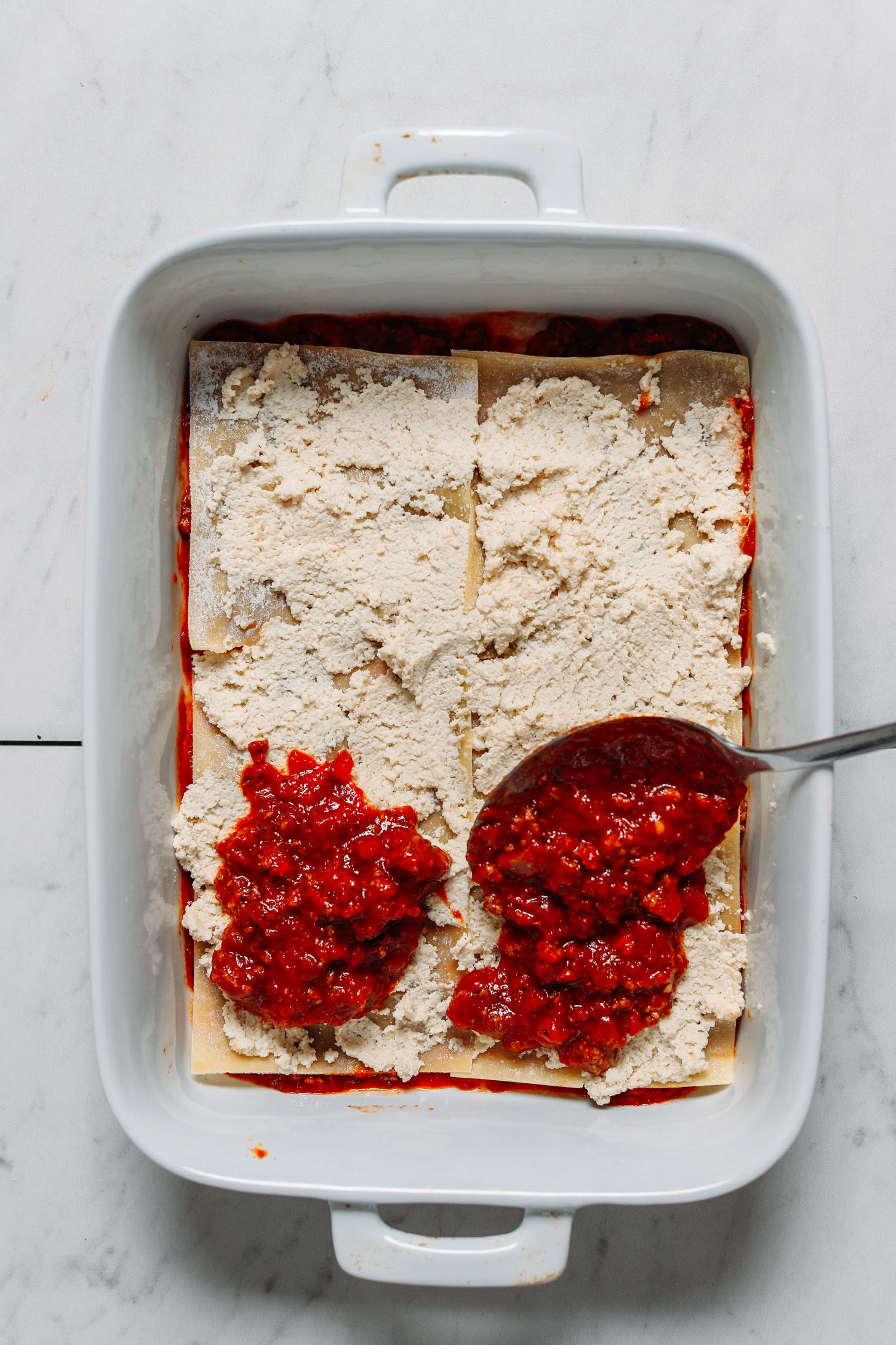 Using a spoon to spread sauce over dairy-free cheese and gluten-free noodles for making lasagna