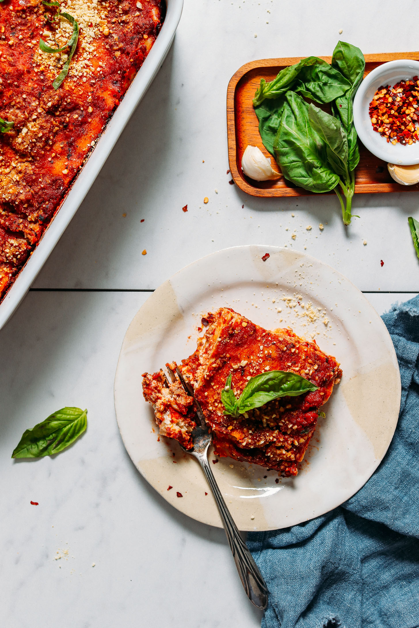 Plate with a slice of lasagna beside fresh basil, garlic, red chili flake, and a pan of the rest of the batch
