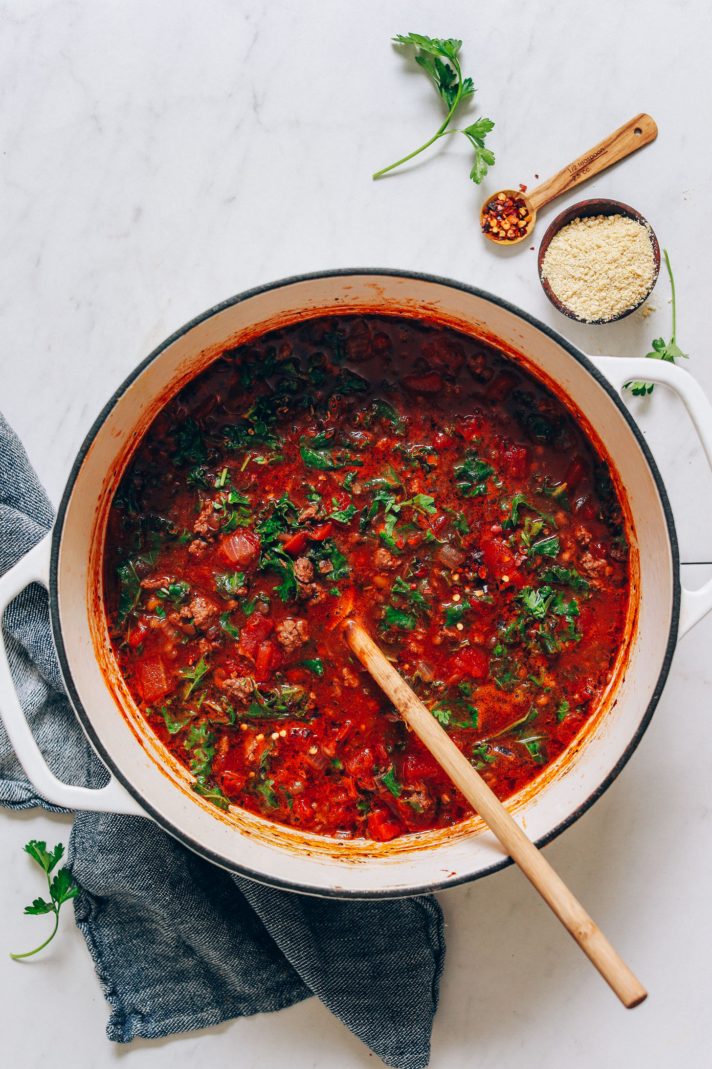 Big pot of Tuscan-Style Beef & Lentil Soup next to fresh parsley, red pepper flake, and cashew parmesan.