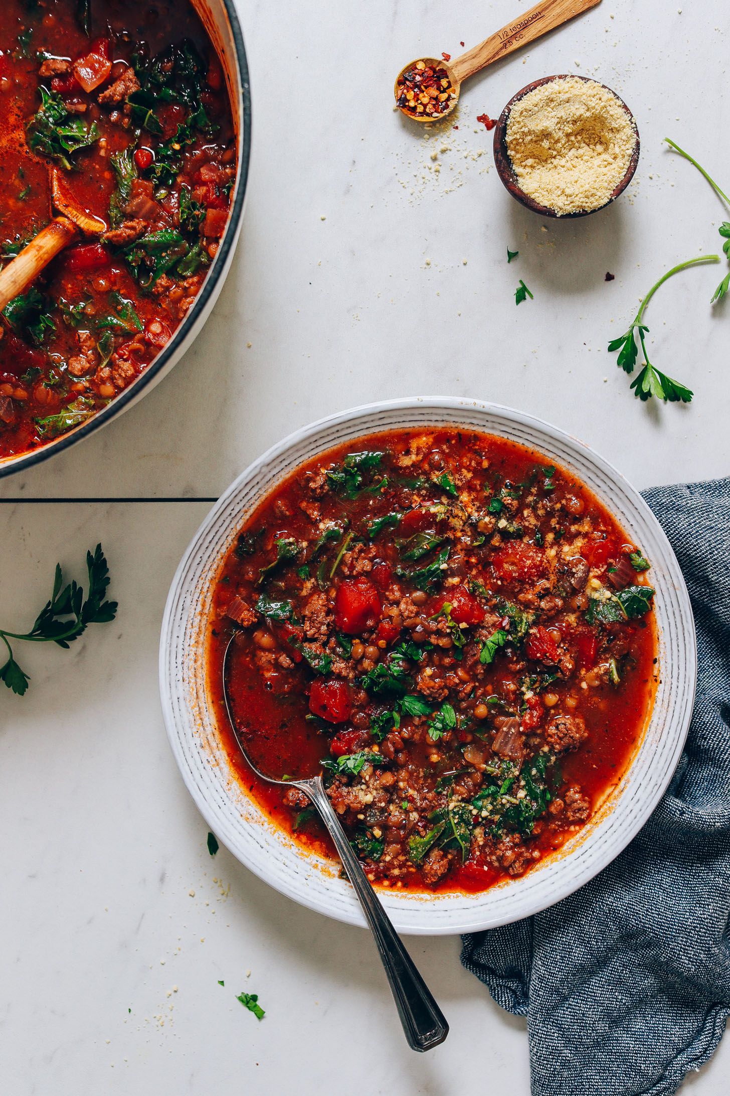 Bowl and pot of Tuscan-Style Beef & Lentil Soup next to vegan parmesan and red pepper flakes for serving