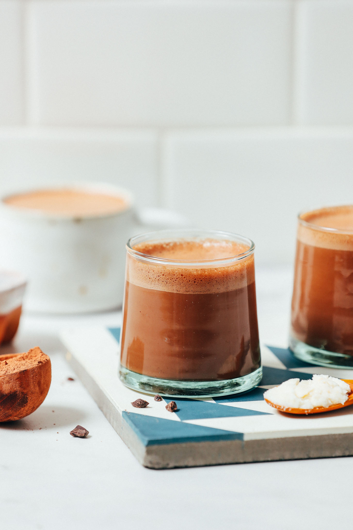 Side view of two glasses and a mug of low sugar Adaptogenic Hot Chocolate beside ingredients used to make it