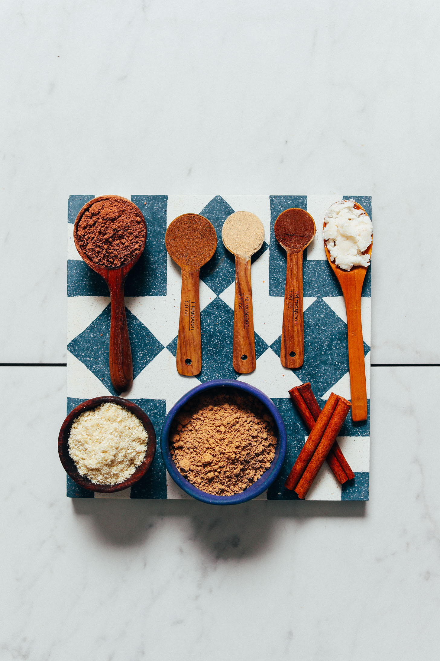 Spoonfuls and bowls of cacao powder, Ashwagandha, reishi mushroom powder, maca, coconut butter, and cinnamon sticks for making our hot chocolate recipe with adaptogens