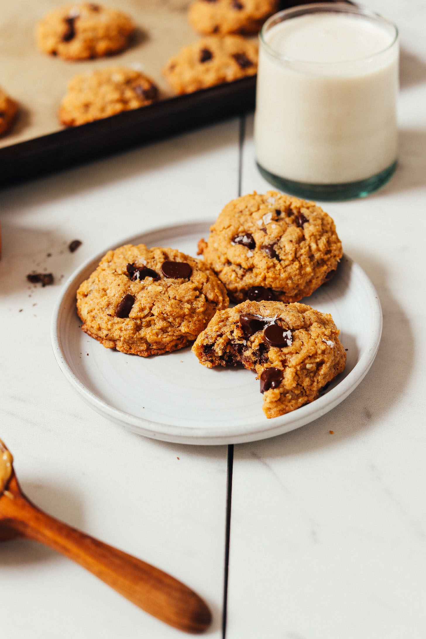 Plate of gluten-free 1-Bowl Peanut Butter Chocolate Chip Cookies next to almond milk and a baking sheet with more cookies