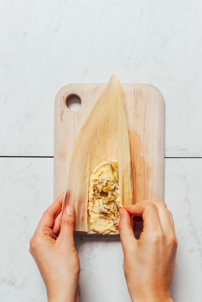 Overhead image of a corn husk on a cutting board with masa and green chile chicken filling, and two hands wrapping