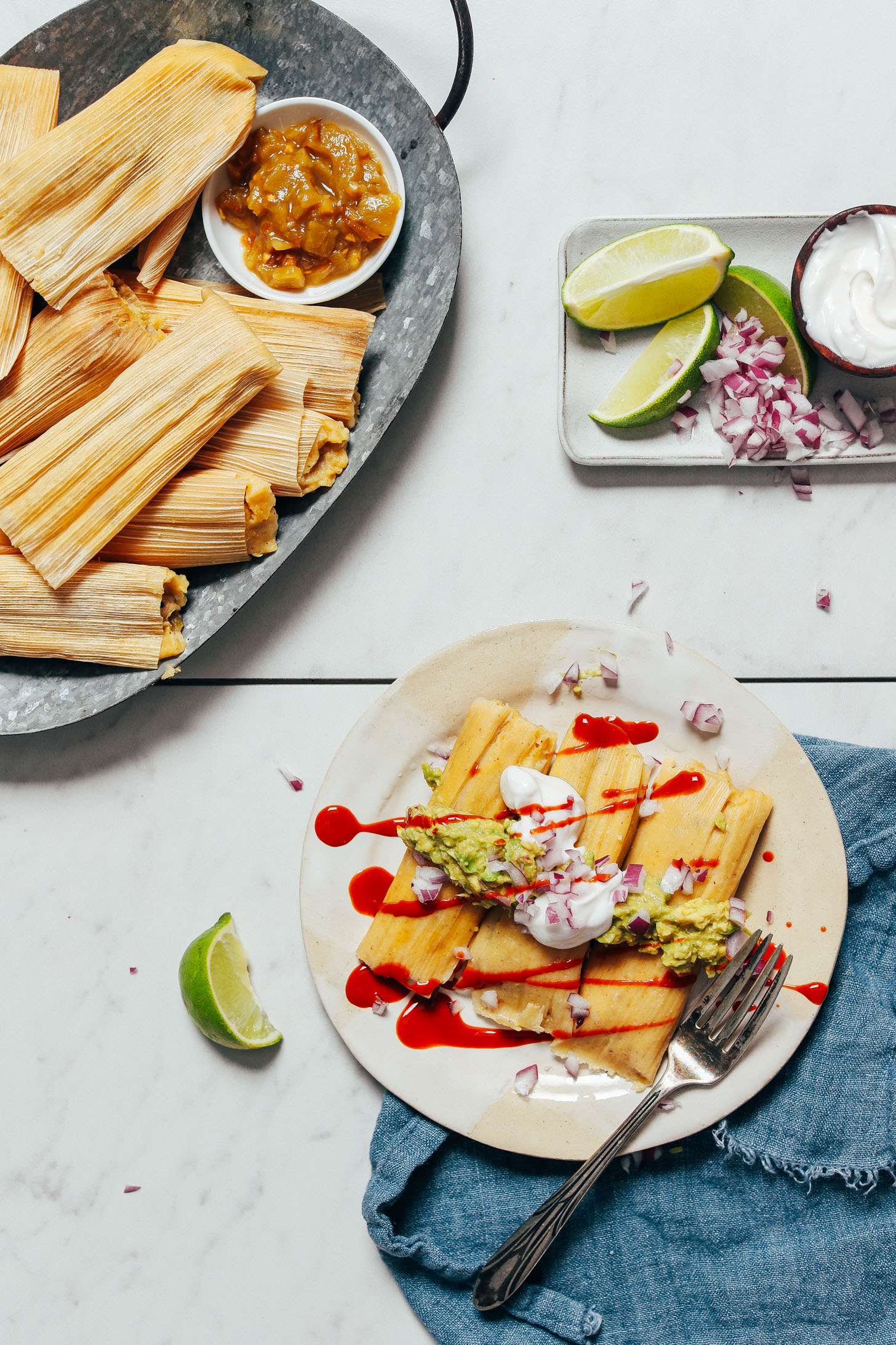 Platter of tamales next to a plate of Green Chile Chicken Tamales topped with guacamole, coconut yogurt, red onion, and hot sauce