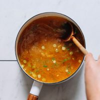 Using a wooden spoon to stir a pot of Chicken Bone Broth with green onion and seasonings