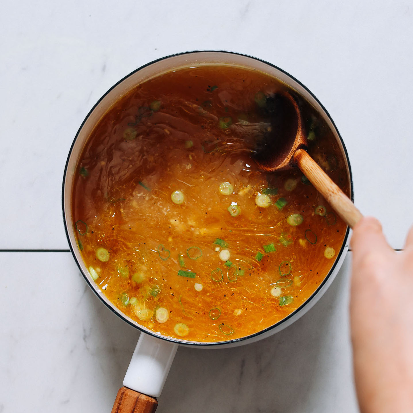 Using a wooden spoon to stir a pot of Chicken Bone Broth with green onion and seasonings