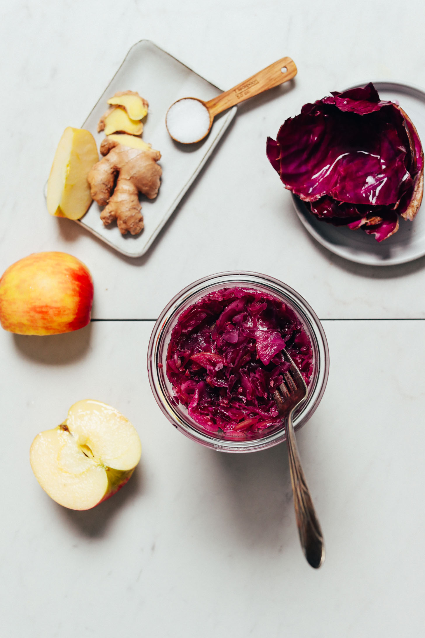 Tall jar of homemade Sauerkraut next to cabbage, apple, ginger, and salt used to make it