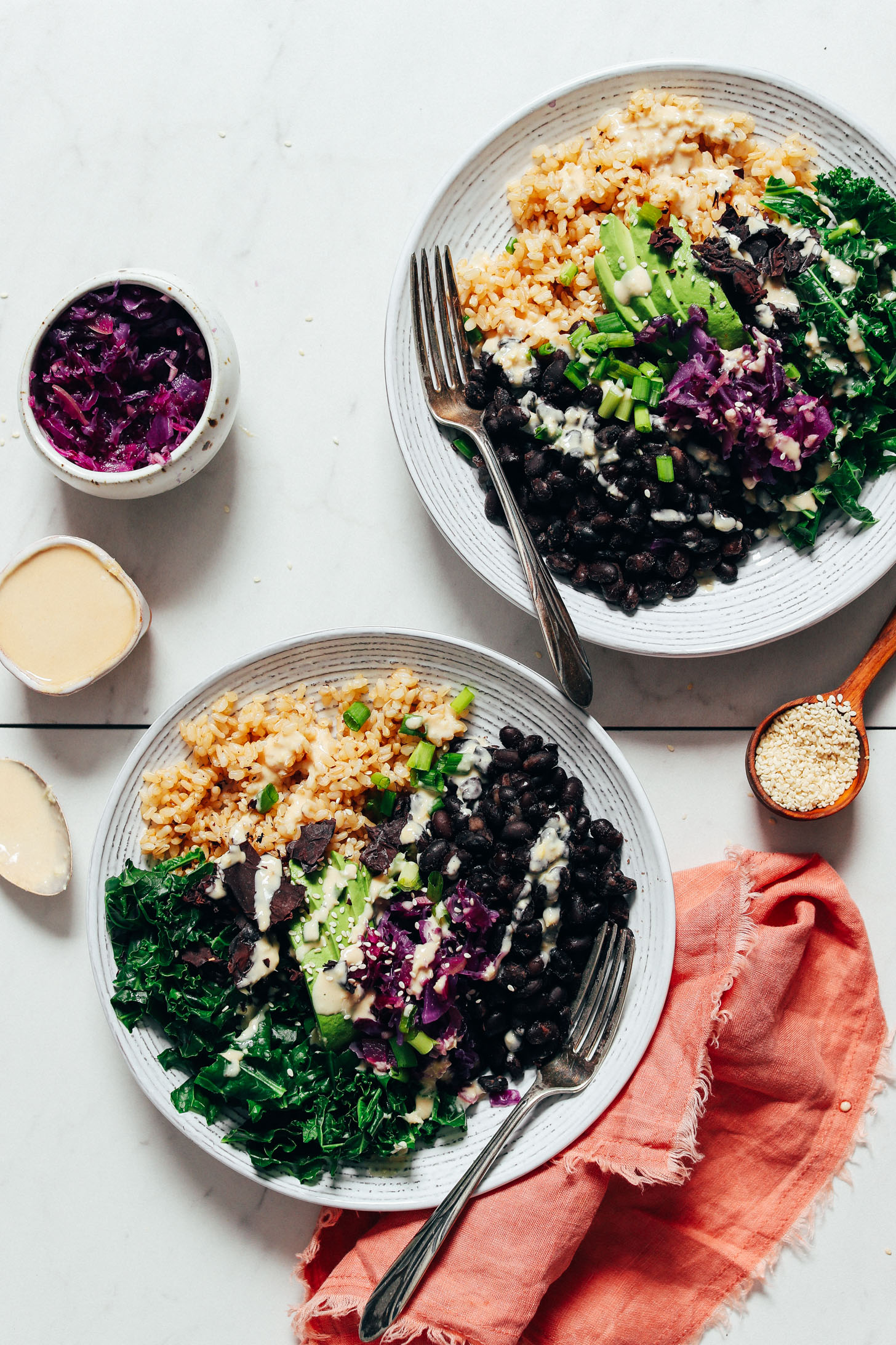 Two Black Bean Buddha Bowls beside sesame seeds, Lemon Ginger Tahini Dressing, and Apple Cabbage Kraut