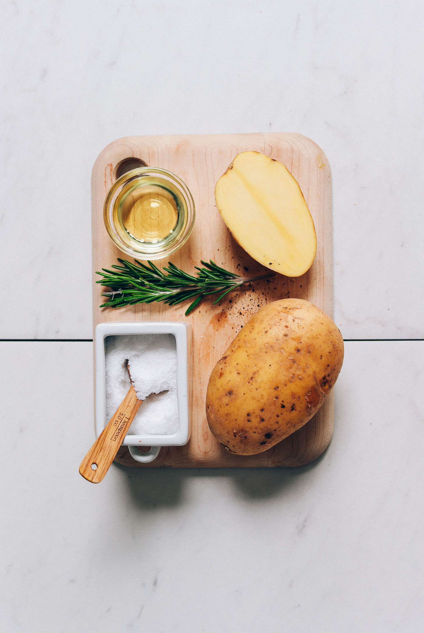 Small cutting board with gold potatoes, avocado oil, rosemary, and salt for making Oven Roasted Potatoes