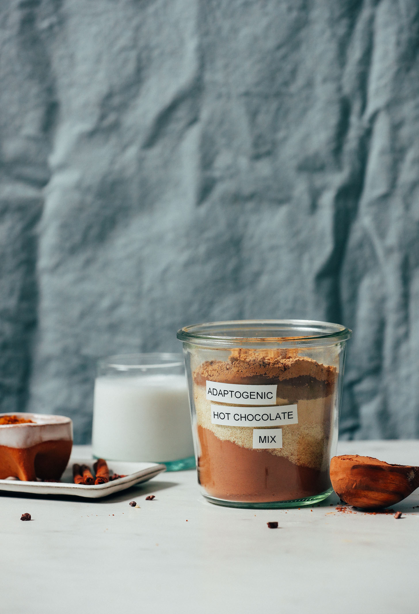 Cinnamon sticks, cacao powder, almond milk, and coconut sugar beside a jar of Adaptogenic Hot Chocolate Mix