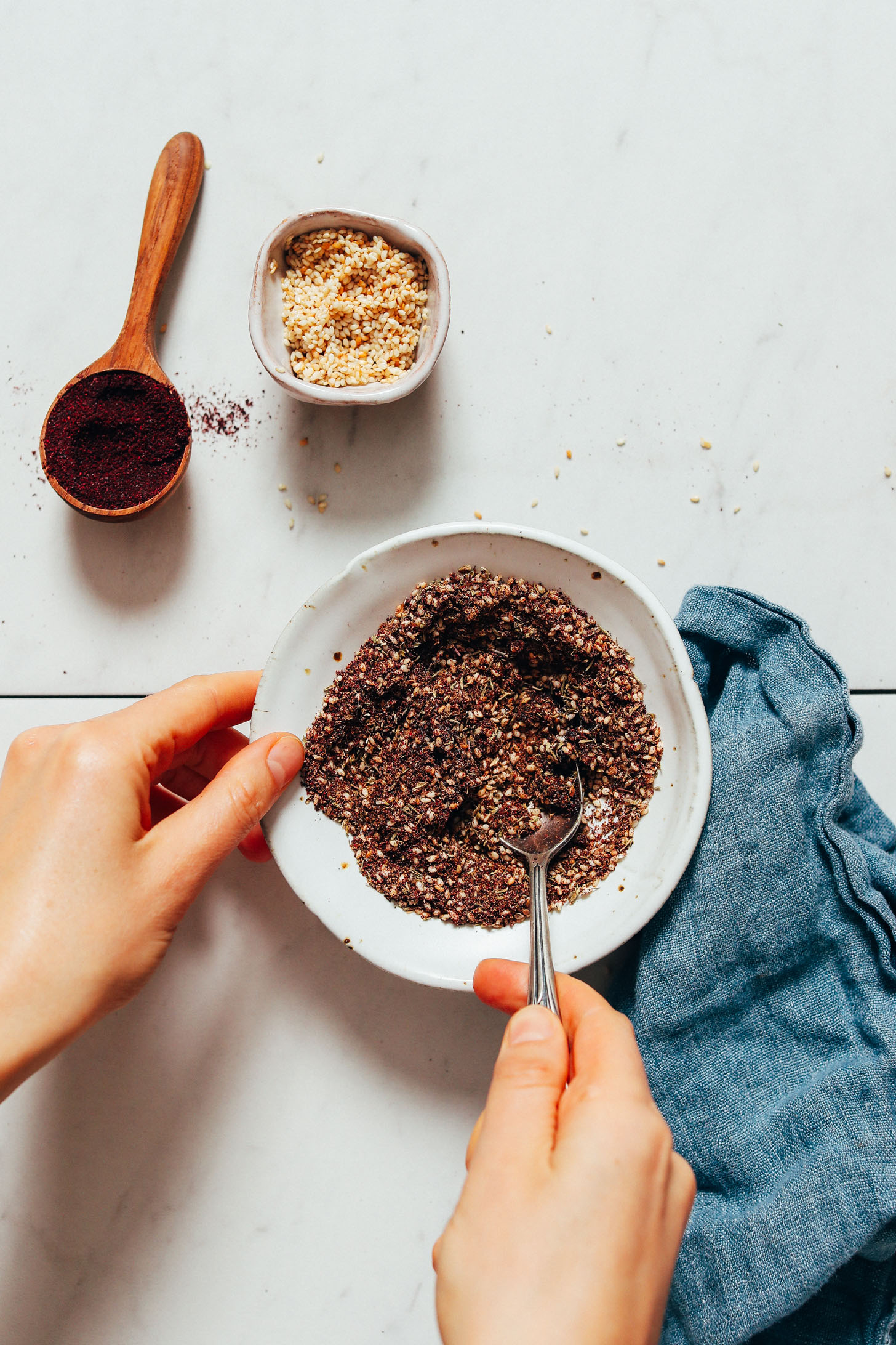 Spoon in a bowl of homemade za'atar seasoning next to a spoonful of sumac and bowl of toasted sesame seeds