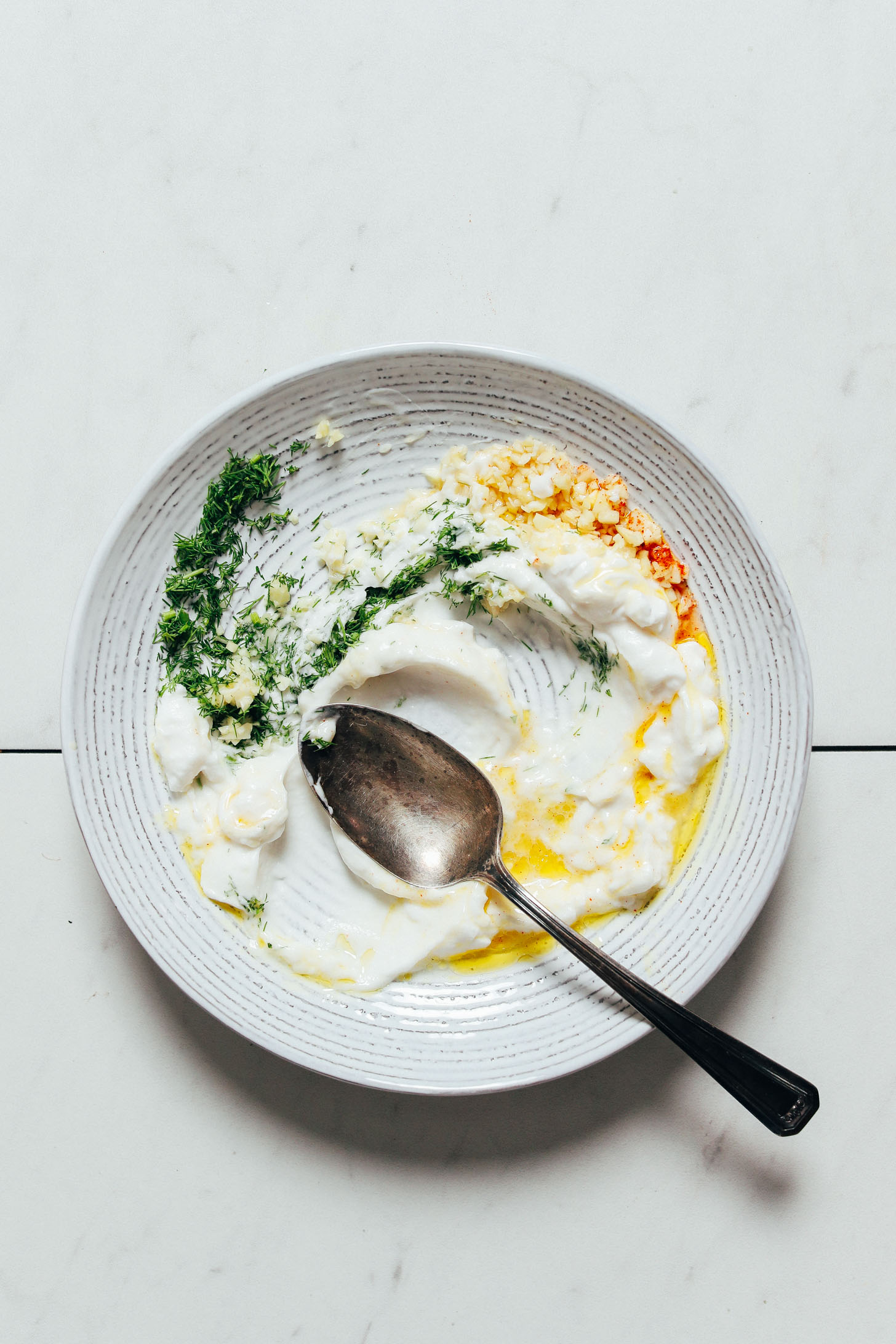 Spoon resting in a bowl filled with vegan yogurt, olive oil, lemon juice, cayenne, and dill