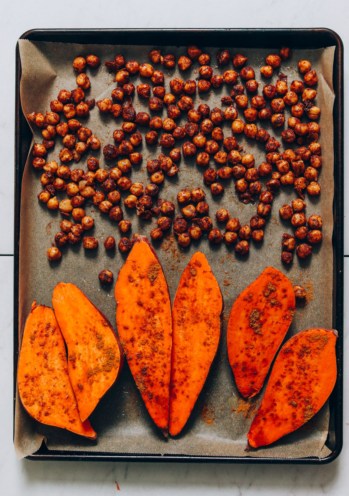 Parchment-lined baking sheet of sweet potatoes and chickpeas topped with tandoori masala seasoning
