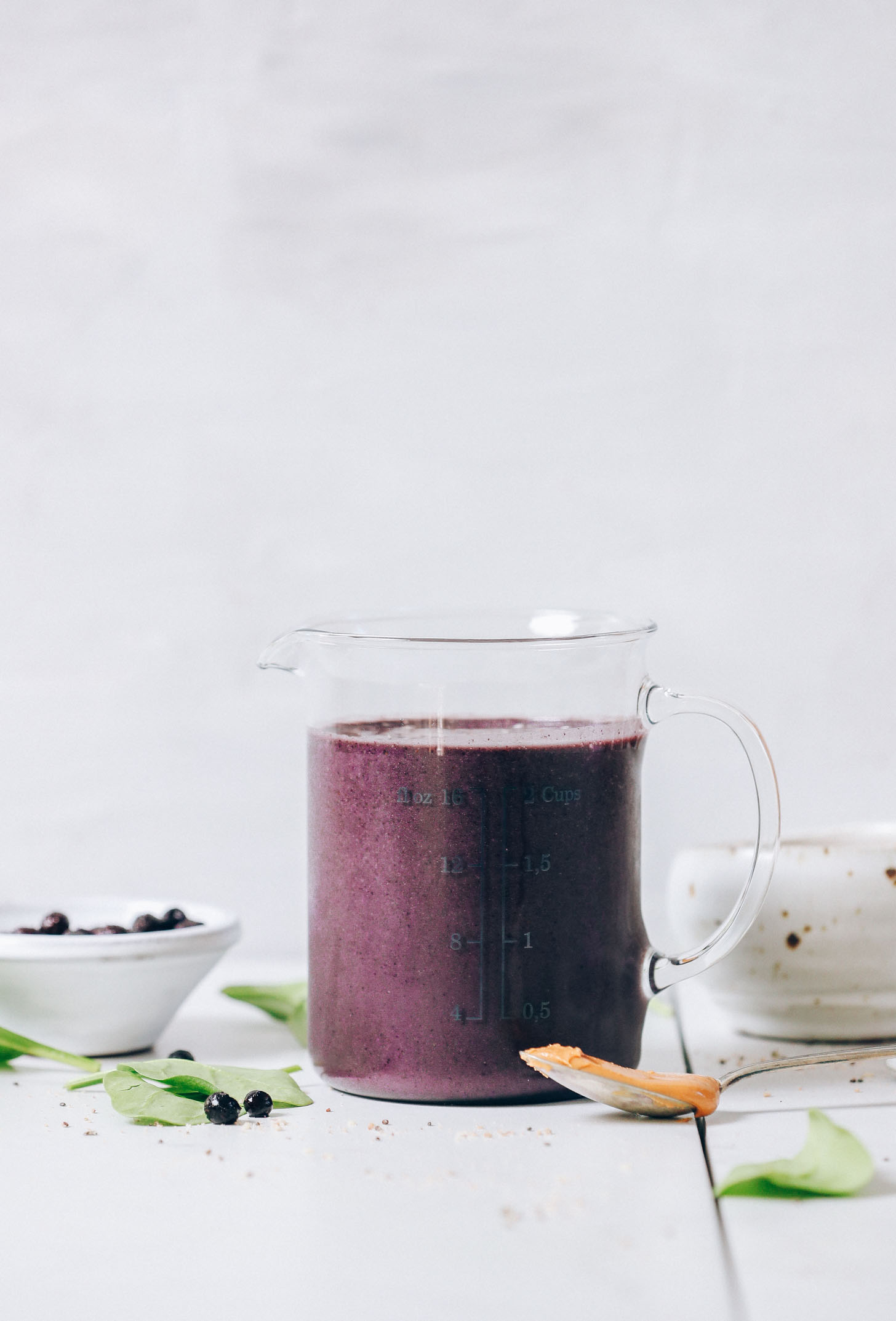 Blueberry spinach peanut butter smoothie in a glass measuring cup with a spoonful of peanut butter, spinach, and blueberries around it.